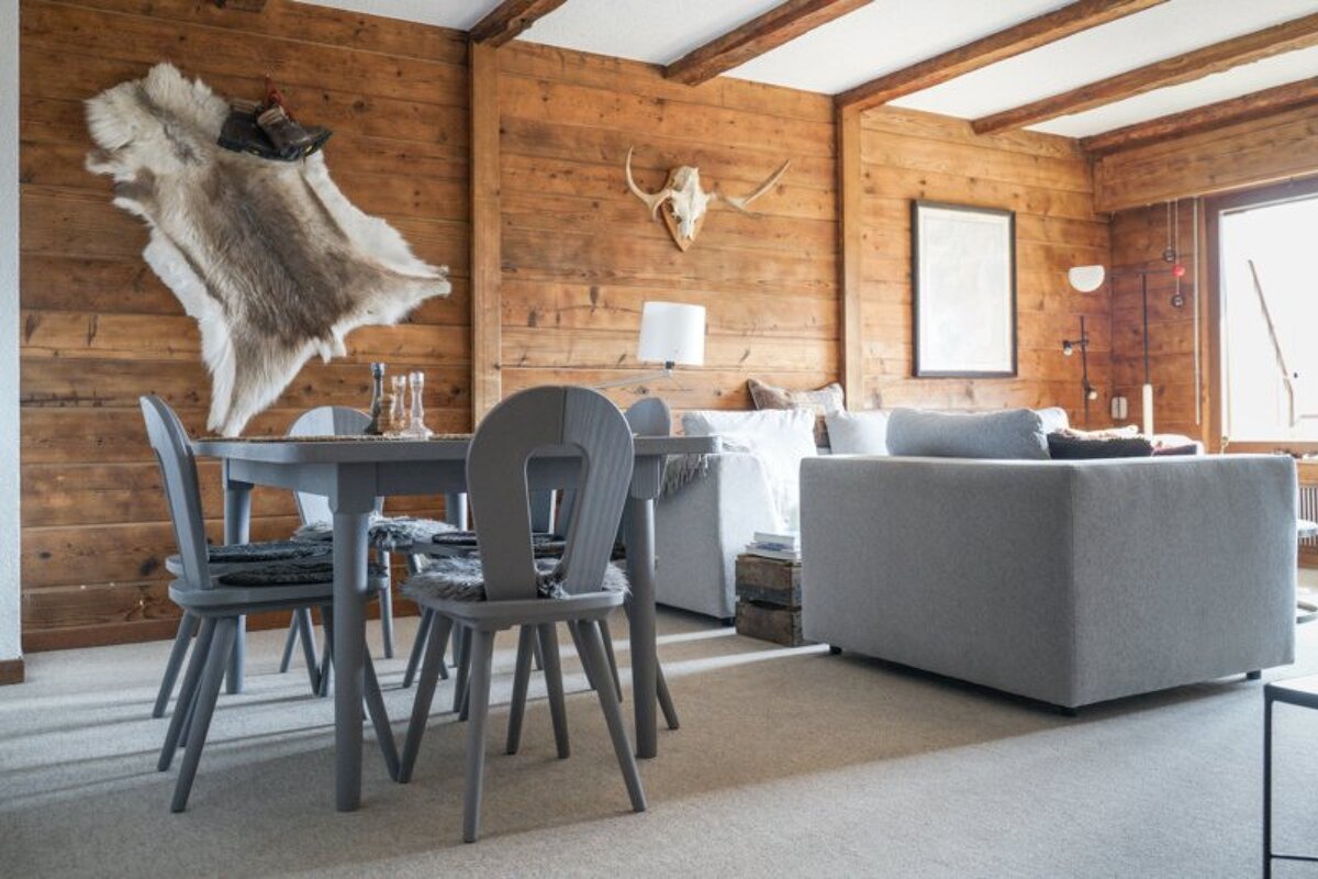 Cozy cabin interior featuring wood-paneled walls, an animal hide wall hanging, a grey dining table, and a grey sofa.