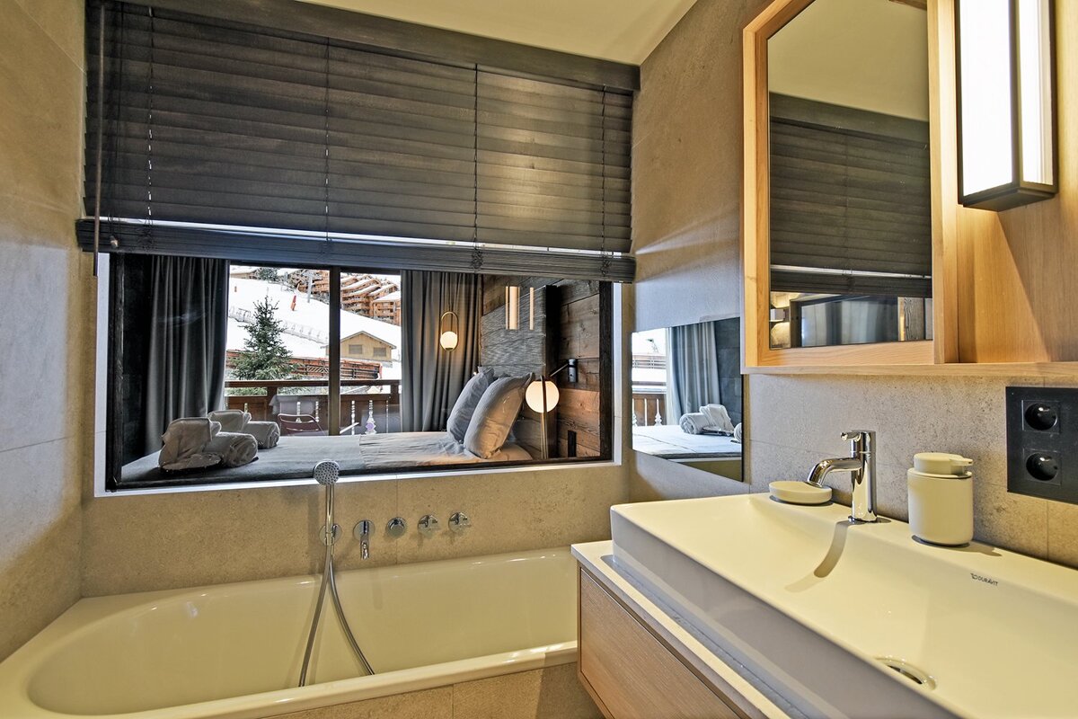 A chic bathroom featuring a tub and vanity next to a large interior window overlooking a cozy bedroom with snowy mountain views.