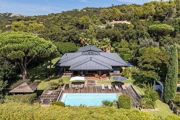 An aerial view of a luxurious modern house with a grey roof, a sparkling swimming pool, and a wooden deck, nestled in a lush, green, tree-covered hillside under a clear sky.