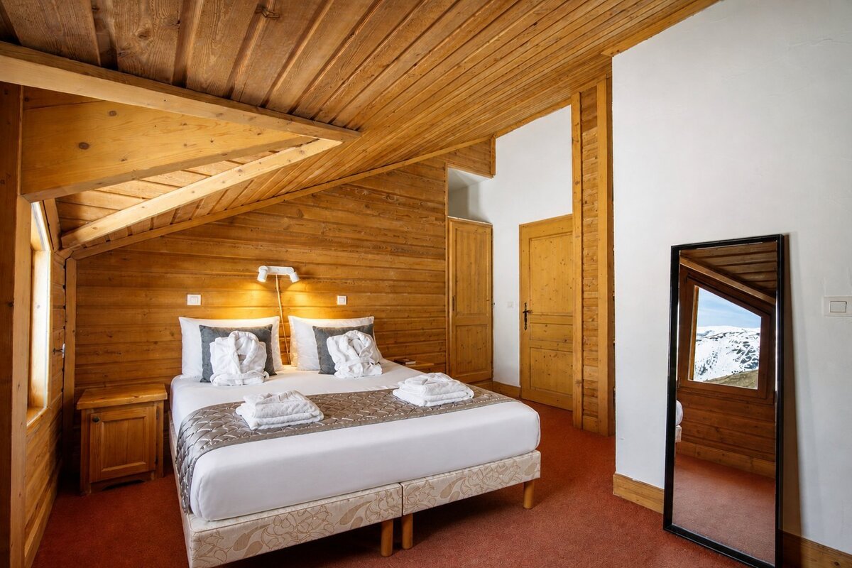 Warm, rustic chalet bedroom with wooden walls and ceiling, a neat double bed, and a mirror reflecting snowy mountains.