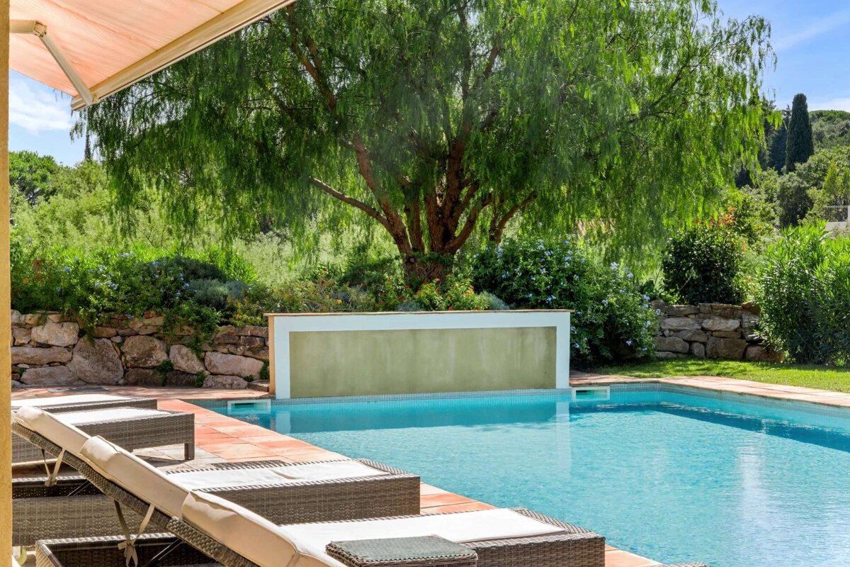 A tranquil outdoor swimming pool with lounge chairs, surrounded by lush greenery, a large tree, and a feature wall, partly shaded by an awning.