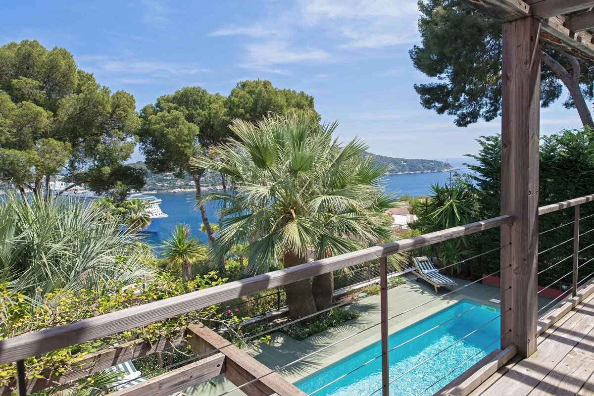 An elevated view from a wooden deck of a blue pool, green palms, and a sparkling bay with a cruise ship under a clear sky.