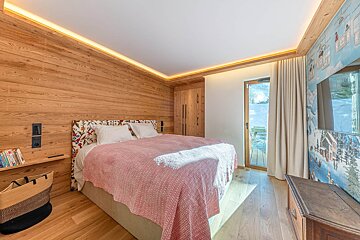 A rustic, wood-paneled bedroom with a pink bedspread, floral headboard, ski-themed wall art/TV, and a balcony door offering outdoor views.