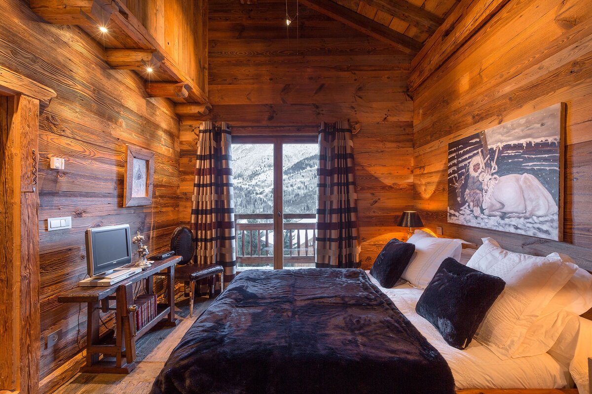 A cozy, rustic wooden chalet bedroom with a bed, small TV, and a picturesque view of snow-covered mountains from the window.
