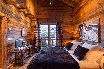 A cozy, rustic wooden bedroom with a plush bed, window overlooking snowy mountains, and a TV. Artwork hangs above the bed.