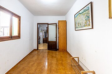 A narrow hallway with white walls, brown tiled floor, a window, an open wooden door, a cabinet, a framed map, and a shoe rack.