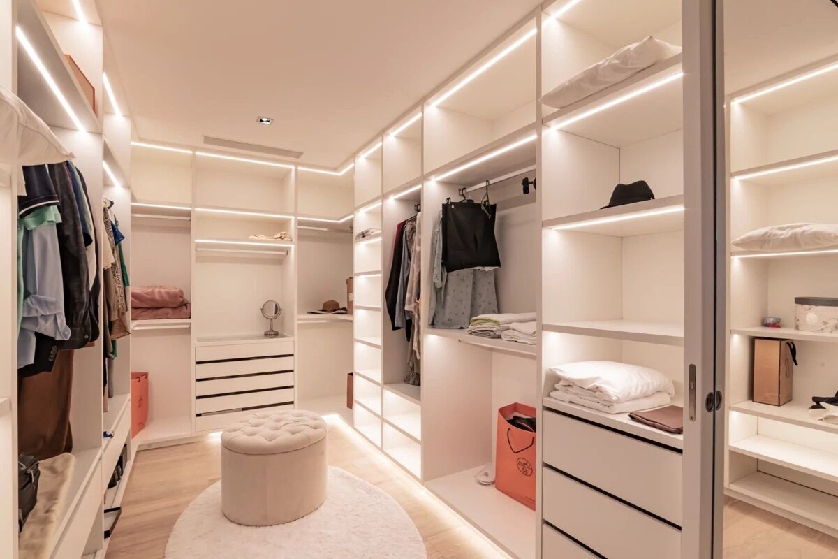 A modern walk-in closet with white shelves, built-in LED lighting, a round vanity stool, and organized clothes and accessories.