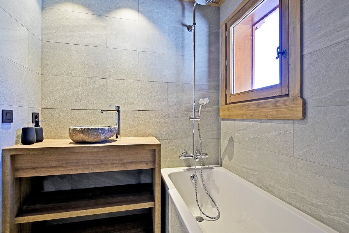 Modern bathroom featuring a stone sink on a wooden vanity, a white bathtub with a shower, grey tiled walls, and a wooden window.