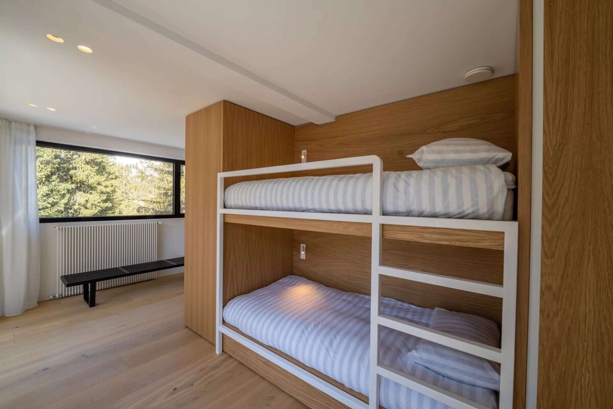 A modern bedroom features built-in light wood bunk beds with striped bedding. A large window overlooks a lush forest, complementing the clean design and wood floor.