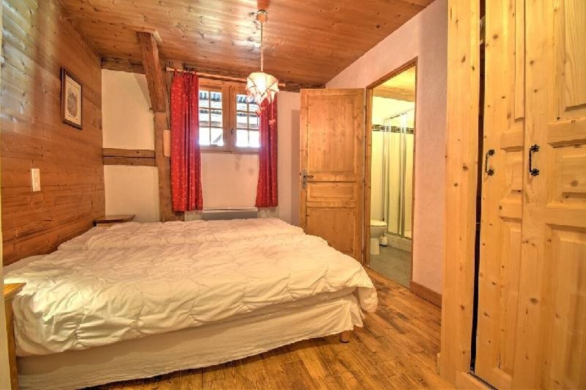 A cozy, all-wood bedroom with a large bed, red-curtained window, and an open door revealing an ensuite bathroom with a toilet and shower.