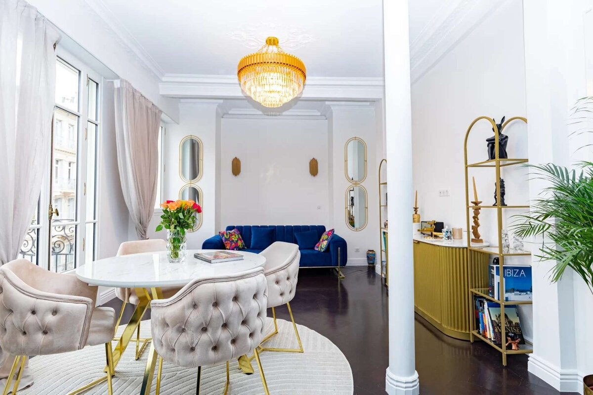 A chic, opulent living space featuring a white dining table with plush chairs, a blue sofa, gold chandelier, and elegant gold accents.