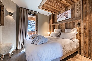 Cozy, wood-paneled chalet bedroom with a large bed, white linens, fox artwork, and a balcony view of a snowy village.
