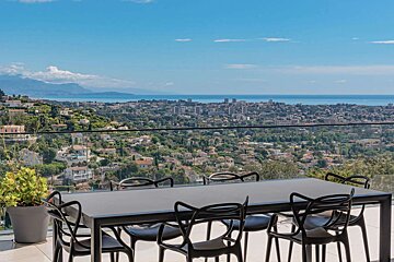 A modern outdoor dining set overlooks a panoramic view of a coastal city nestled on green hills, with the blue sea under a clear sky.