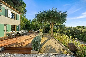 Sunny Mediterranean house with green shutters, a wooden deck patio, outdoor dining area, and a lush green garden under a clear blue sky.