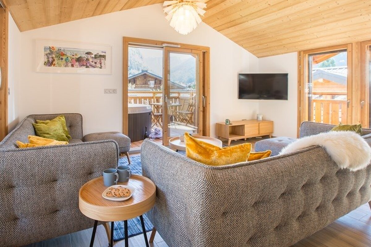 Cozy mountain chalet living room featuring grey tufted sofas with yellow pillows, wooden ceilings, and a scenic balcony view.