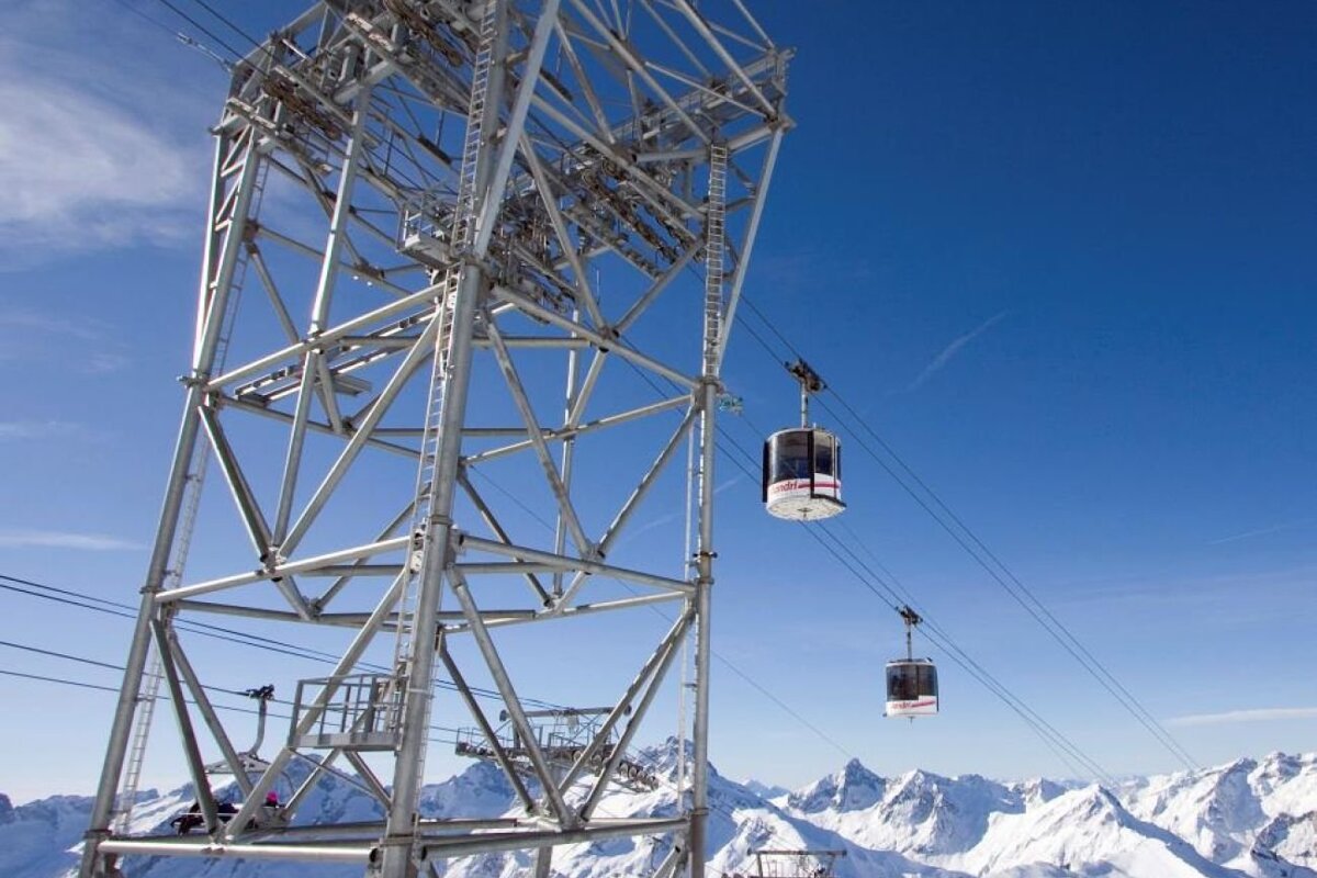 Cable cars suspended from a massive metal tower glide over snowy mountain peaks under a bright blue sky.