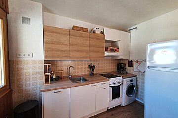 A bright kitchen with wood-grain upper cabinets, white lower cabinets, and a patterned tiled backsplash. It includes a sink, stove, washing machine, and fridge.
