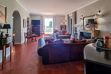 A spacious living room with warm terracotta floors, burgundy sofas, a fireplace, and a balcony view, featuring classic decor.