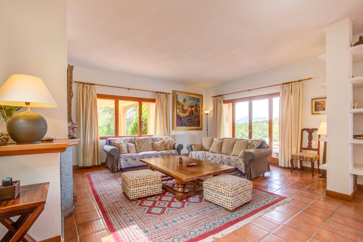 A bright, rustic living room with two comfy sofas, a wooden coffee table, and a patterned rug on terracotta floors. Large windows overlook greenery.