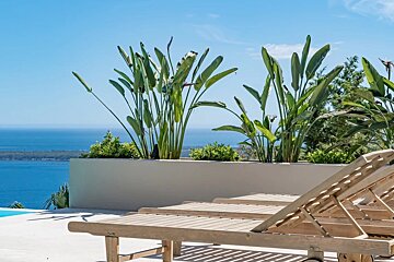 Wooden lounge chairs on a sunny terrace with lush tropical plants overlooking the calm blue ocean.