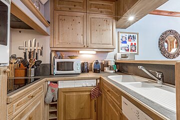 A compact, rustic wooden kitchen featuring light cabinets, a microwave, sink, coffee maker, and various utensils, with homely decor.