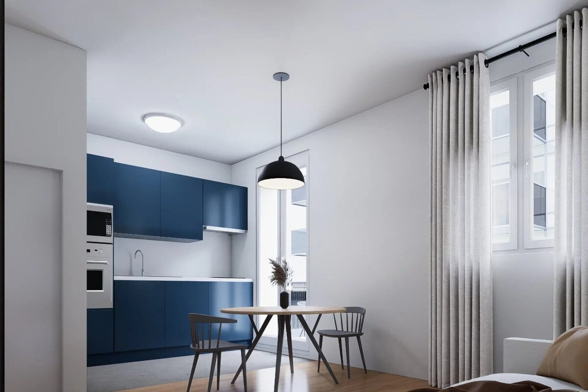 A modern apartment featuring a dark blue kitchen and a small wooden dining table under a black pendant light.