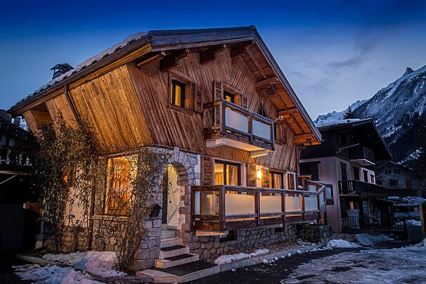 A charming wooden chalet with illuminated windows and snow-dusted grounds, seen at twilight against a dramatic mountain range.