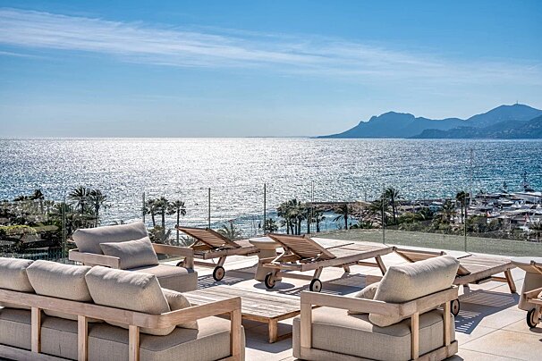 A sunny rooftop patio with lounge chairs and sofas overlooking a shimmering ocean and distant mountains.
