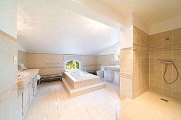 A spacious, light-toned tiled bathroom with an elevated bathtub, walk-in shower, two vanities, and an arched window overlooking greenery.