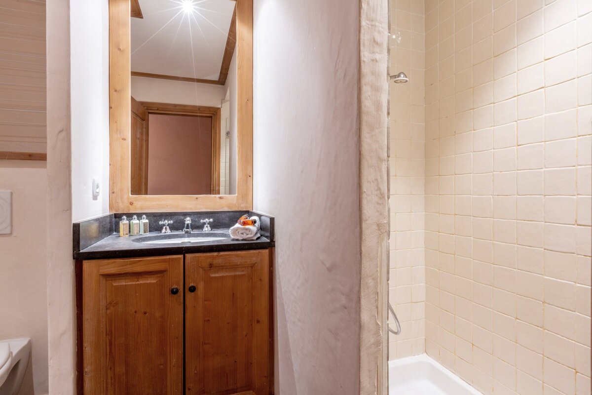 A chalet-style bathroom featuring a wooden mirror over a dark vanity, a cream-tiled shower, and a partial view of a toilet.