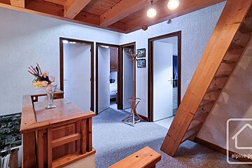 An alpine-style interior features exposed wooden beams, multiple doorways to rooms, a steep wooden staircase, a small bar, and a coat rack.