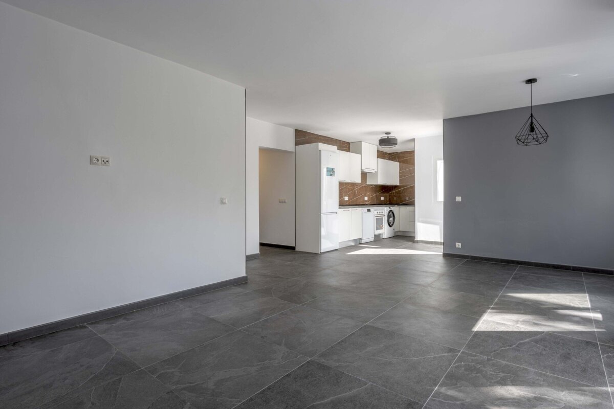An empty, modern open-plan room with grey tiled floors, white and grey walls, and a visible kitchen featuring white cabinets and a brown backsplash.