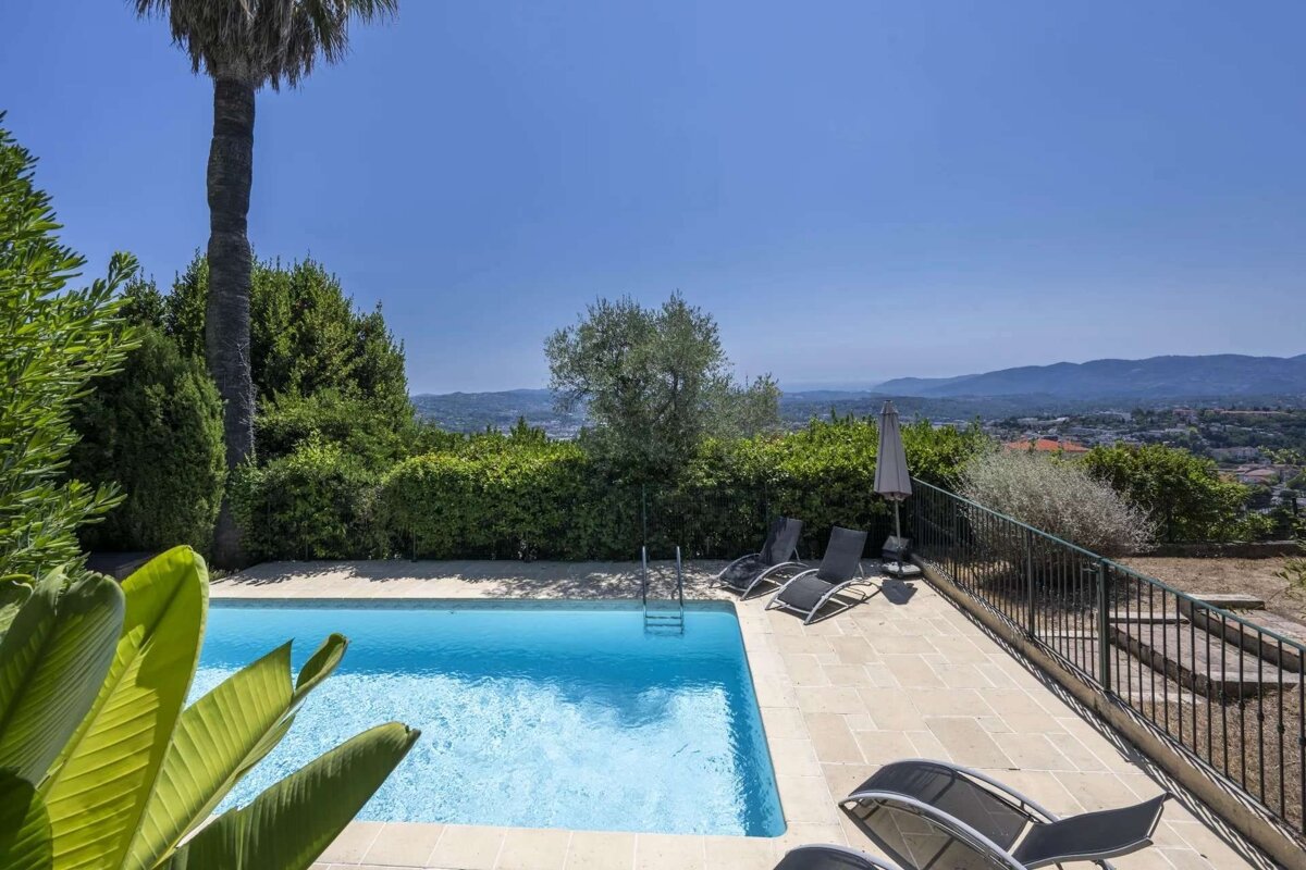 A sun-drenched swimming pool with lounge chairs and palm trees, set amidst lush greenery, offering a panoramic view of distant hills and a town.