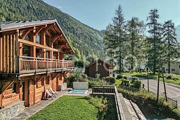 A rustic wooden alpine chalet featuring an outdoor hot tub and green lawn, nestled against a backdrop of pine-covered mountains.