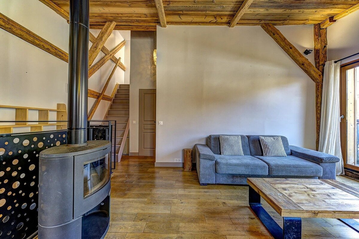A rustic living room featuring exposed wooden beams, a black wood stove, a gray sofa, and a wooden coffee table, with a staircase visible.