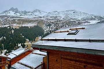 Snow-dusted wooden chalets and pine forests against a backdrop of majestic, snow-capped mountains under an overcast sky.