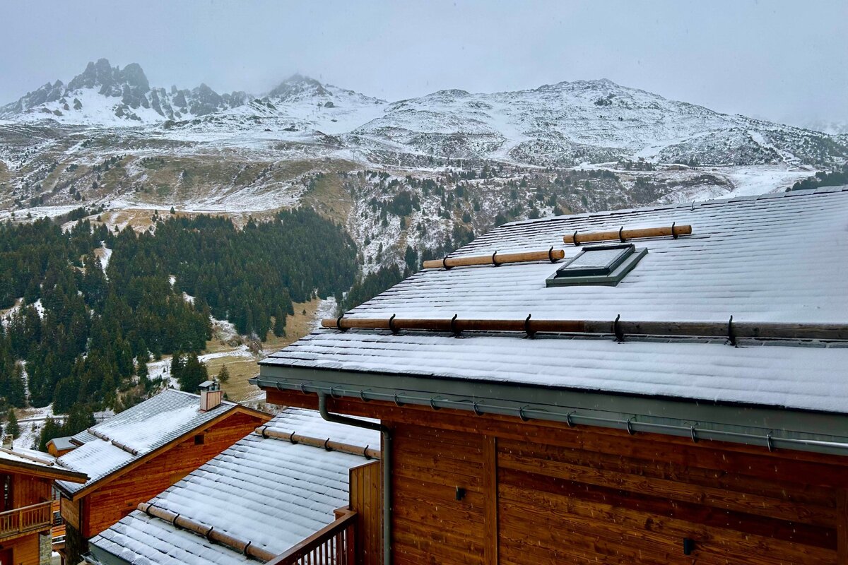 Snow blankets wooden roofs of mountain cabins, with pine forests and rugged, snow-capped peaks rising in the background under a cloudy sky.