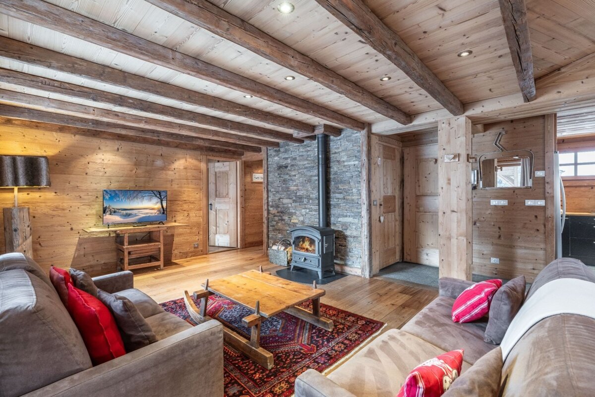 A cozy, rustic living room with warm wood paneling, exposed beams, a lit wood stove, comfy sofas, a wooden coffee table, and a TV.