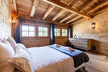 Cozy cabin bedroom with wood-paneled walls, exposed ceiling beams, a neatly made bed, and a wooden dresser.