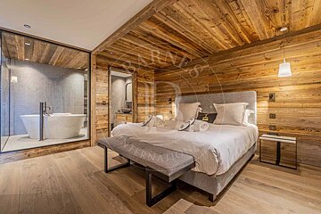 Warm bedroom with wood-paneled walls/ceiling, a large bed, and a modern bathroom with a freestanding tub behind a glass wall.
