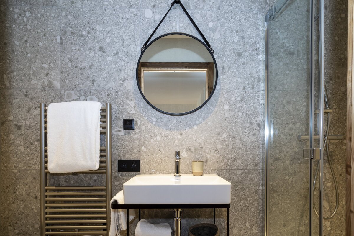 Modern bathroom: grey speckled tiles, round mirror, white sink, towel warmer, and glass shower.