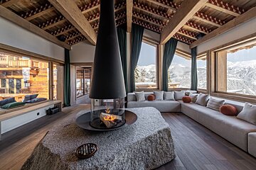Warm, modern chalet interior with a central suspended fireplace, plush seating, and expansive windows showcasing snowy mountain vistas.