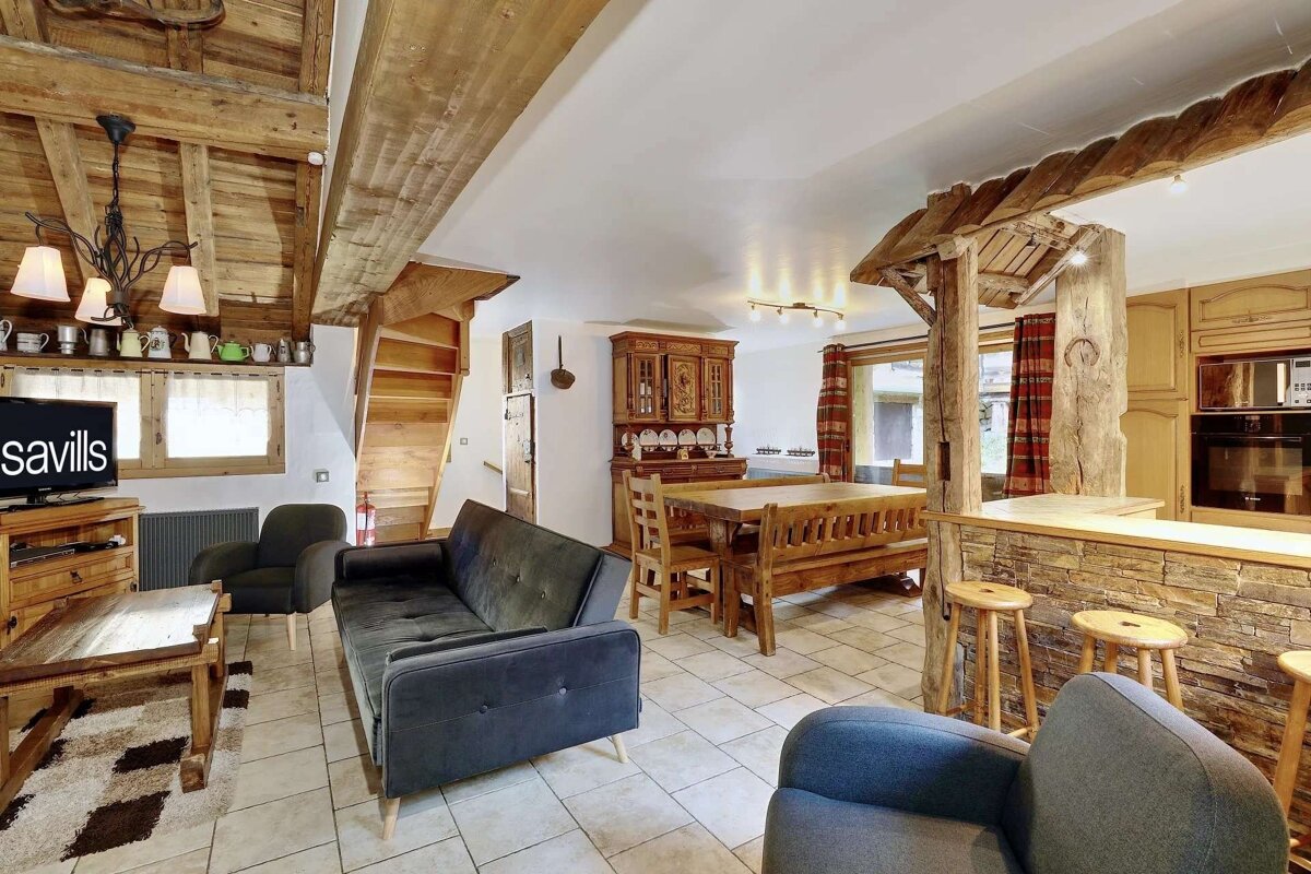 A spacious, rustic open-plan living area with exposed wood beams, a dining table, kitchen island, and comfortable seating on a tiled floor.