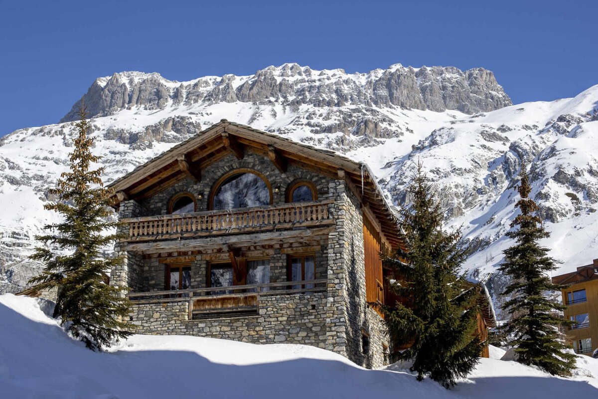 A rustic stone and wood chalet stands in deep snow, framed by green pine trees, against a backdrop of towering, snow-covered mountains.