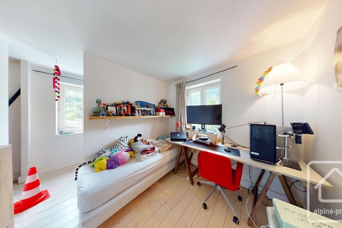 A bright room with white walls, featuring a daybed with plush toys, a desk with a computer and red chair, a shelf, and a window.