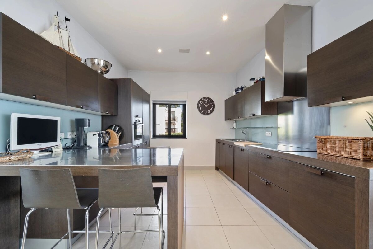 A sleek, modern kitchen featuring dark wooden cabinets, stainless steel appliances, and a central island with bar stools on a tiled floor.
