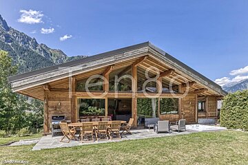 A luxurious wooden chalet with a large patio, outdoor dining, and a hot tub, set against a stunning mountain backdrop.