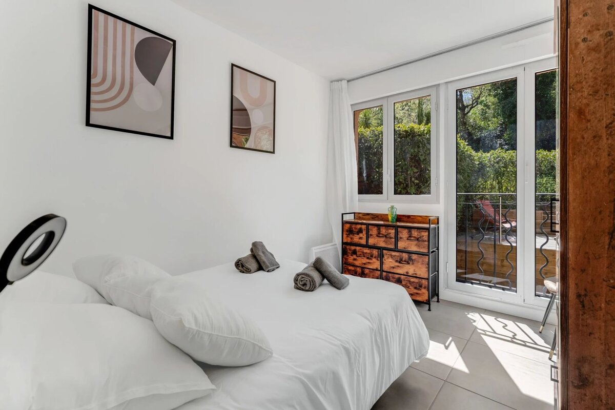 Bright, minimalist bedroom featuring a white bed with rolled towels, abstract art, a wooden dresser, and glass doors to a sunny patio.