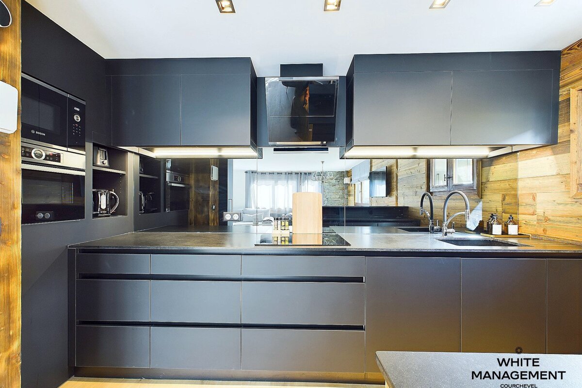 A sleek, dark modern kitchen features integrated appliances, wood accents, and a mirrored backsplash reflecting a bright living area.