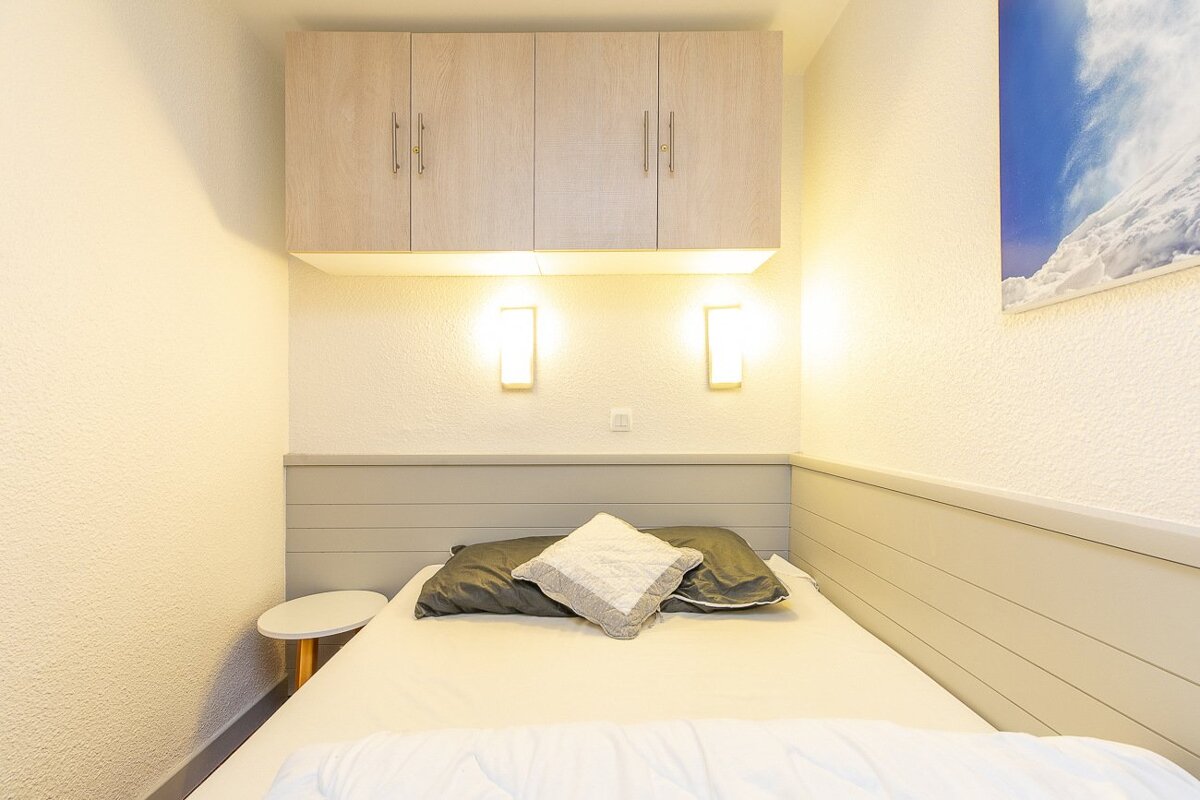 Compact modern bedroom with a bed, overhead wood cabinets, two wall sconces, a side table, and a mountain print on the right wall.